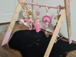 Houten babygym in A-frame stijl, met roze accenten en vijf hangende speeltjes in zachte tinten roze en beige, geplaatst op een houten vloer met een zwart vloerkleed eronder.