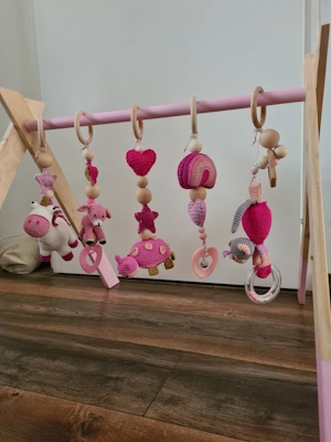 Babygym van hout met roze accenten en vijf handgemaakte hangspeeltjes in roze tinten, waaronder een eenhoorn, hartje, paddenstoel en gehaakte regenboog, geplaatst op een houten vloer.