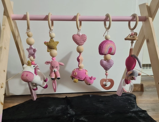 Babygym van hout met roze afwerking, voorzien van vijf gehaakte speeltjes in roze, paars en geel, waaronder een eenhoorn, ster, hartje, regenboog en rammelaar, boven een zwart bontachtig kleed.