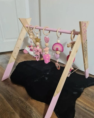 Houten babygym in A-frame stijl, met roze accenten en vijf hangende speeltjes in zachte tinten roze en beige, geplaatst op een houten vloer met een zwart vloerkleed eronder.